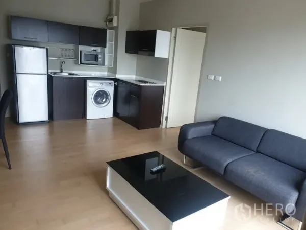 Condominium for rent in Watthana, Bangkok - Open-plan kitchen with dark cabinets, refrigerator, microwave and in-unit washing machine beside the living area.