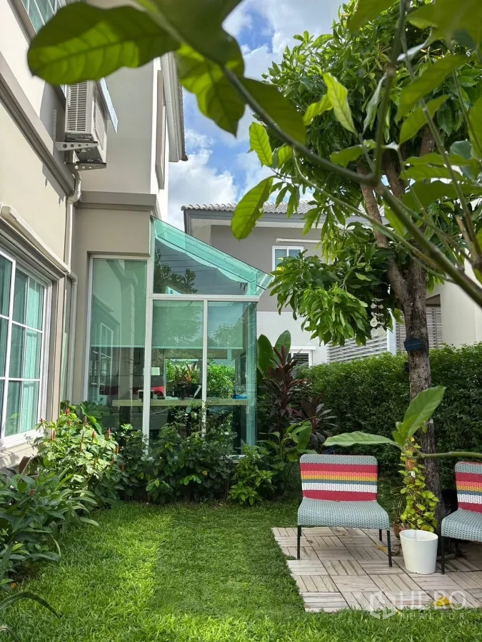 独立式住宅 出租在Prawet, Bangkok - 葱郁花园，带玻璃阳光房、树木与彩色户外椅。