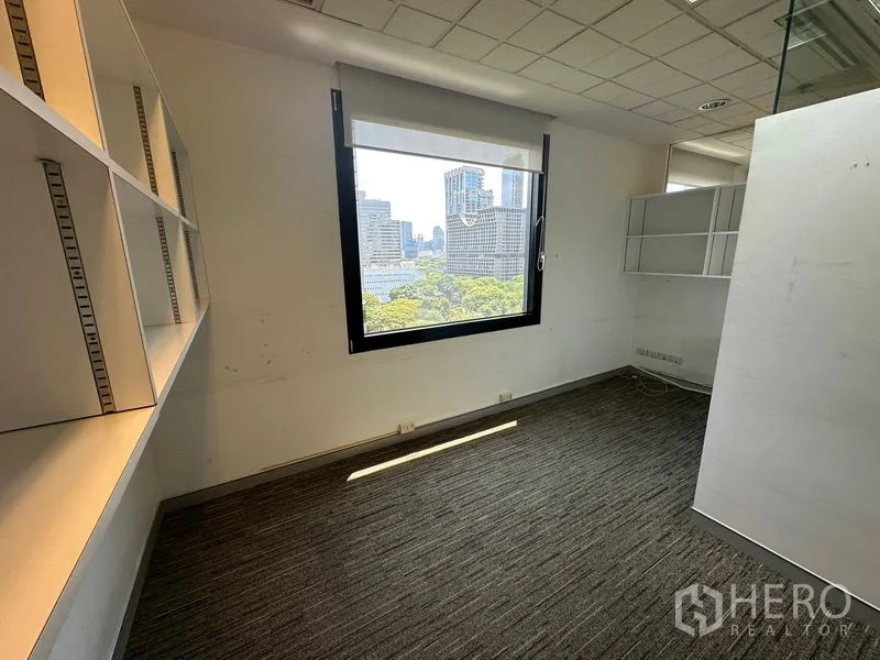 Office Space for rent - Compact workspace with shelving and a large window with roller blind overlooking treetops and high-rise buildings.