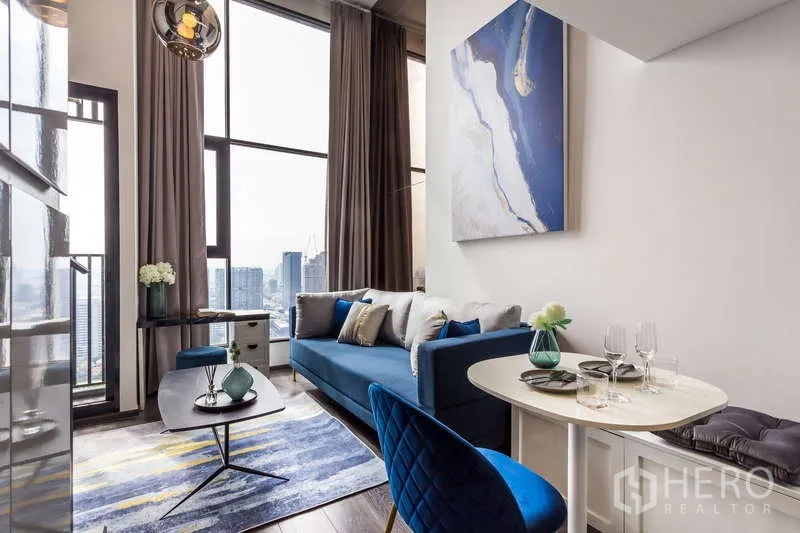 Condominium for rent - Bright living room with blue sofa, dining nook, and floor-to-ceiling windows overlooking the Bangkok skyline.
