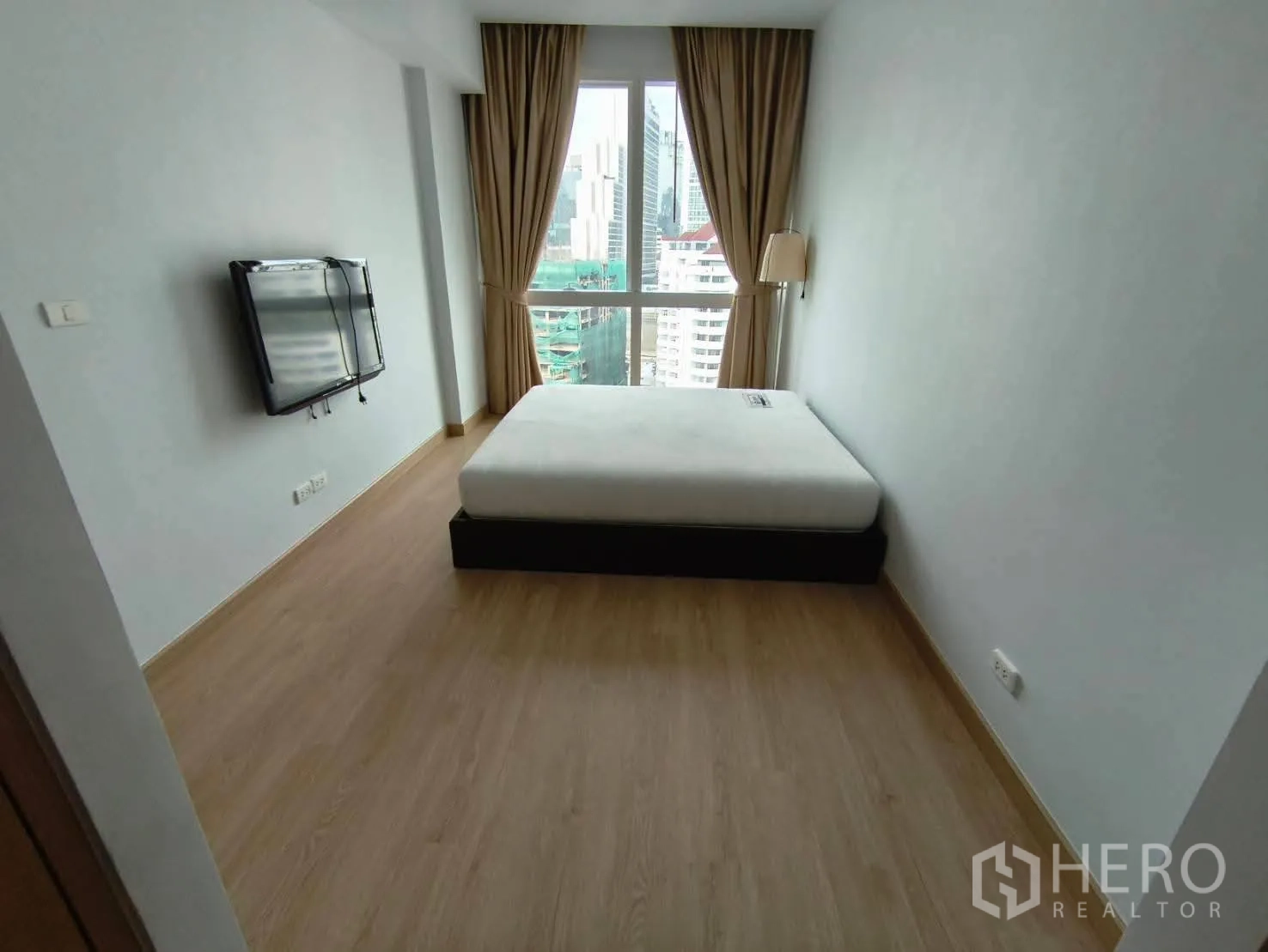 Condominium for rent in Khlong Toei, Bangkok - Master bedroom with city-view window, double bed, wall-mounted TV, and beige curtains.