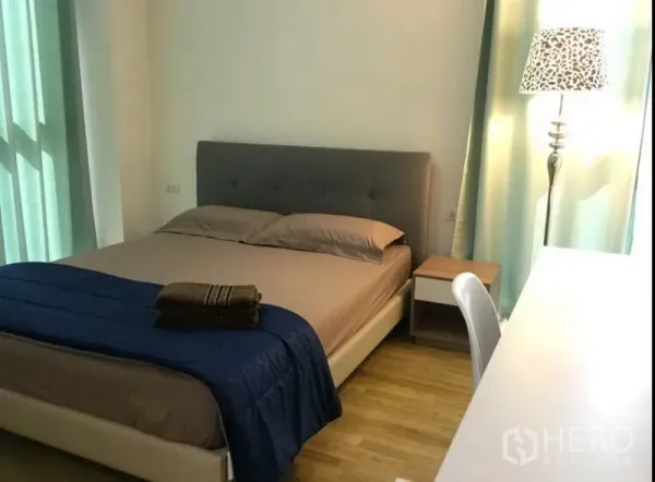 Condominium for rent in Phra Khanong, Bangkok - Master bedroom with upholstered double bed, bedside table, desk, floor lamp and soft green curtains over wood flooring.