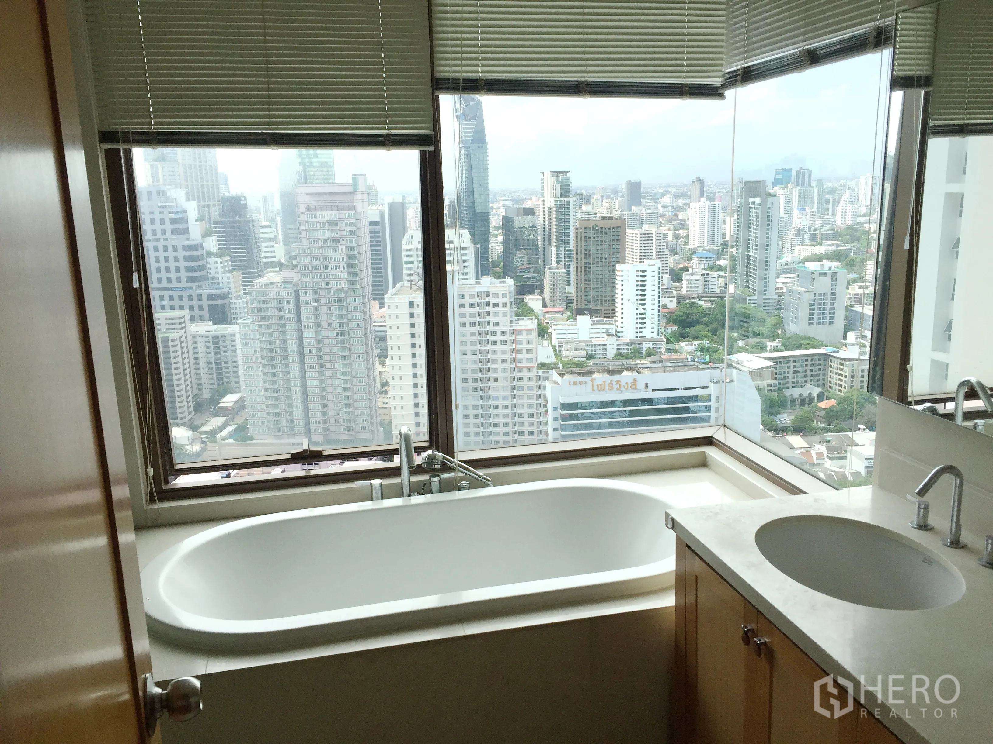Condominium for rent in Khlong Toei, Bangkok - Bathroom with soaking tub beside corner windows overlooking the Bangkok skyline and a stone-top vanity.