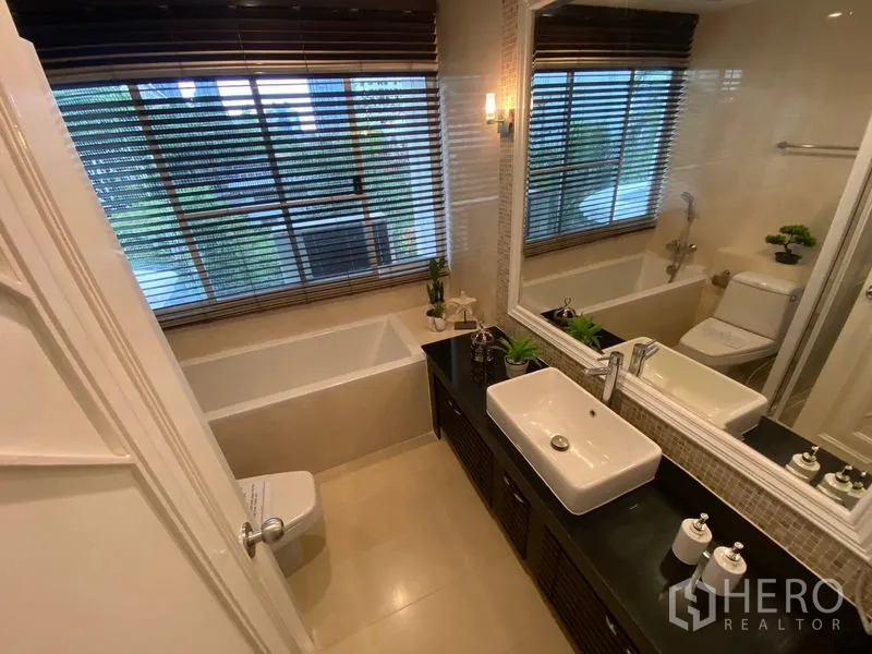 Apartment for rent in Watthana, Bangkok - Bathroom with bathtub, wide mirror and vanity, overlooking greenery through the window.