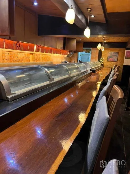 Shophouse for rent in Khlong Toei, Bangkok - Warm-lit wooden sushi counter with refrigerated display cases and high chairs inside the shophouse.
