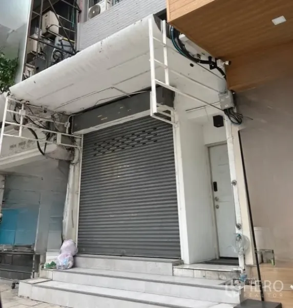 零售空间 出租在Watthana, Bangkok - 一层店面外观，卷闸门关闭，白色立面与台阶，临阿索克繁华街道。
