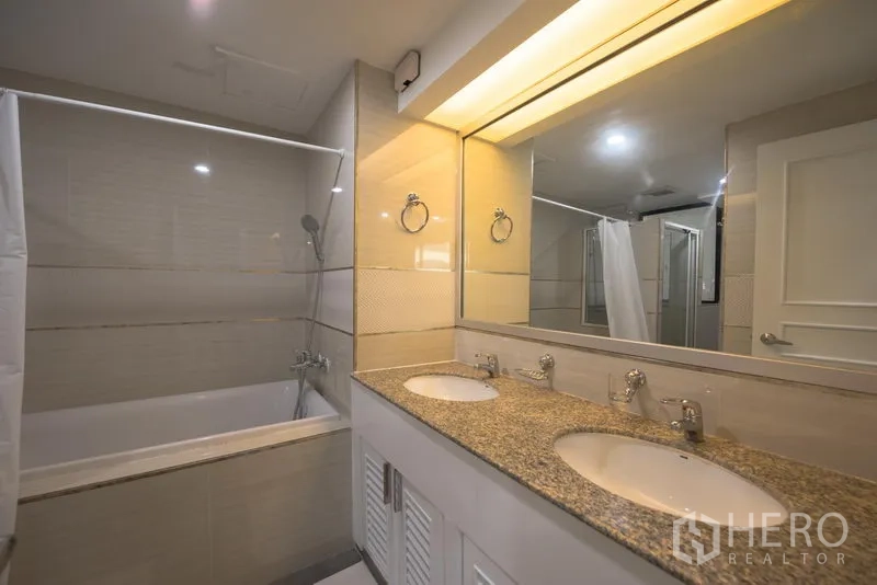 Condominium for rent in Khlong Toei, Bangkok - Bathroom with double sinks on a granite vanity, large mirror and bathtub with shower.