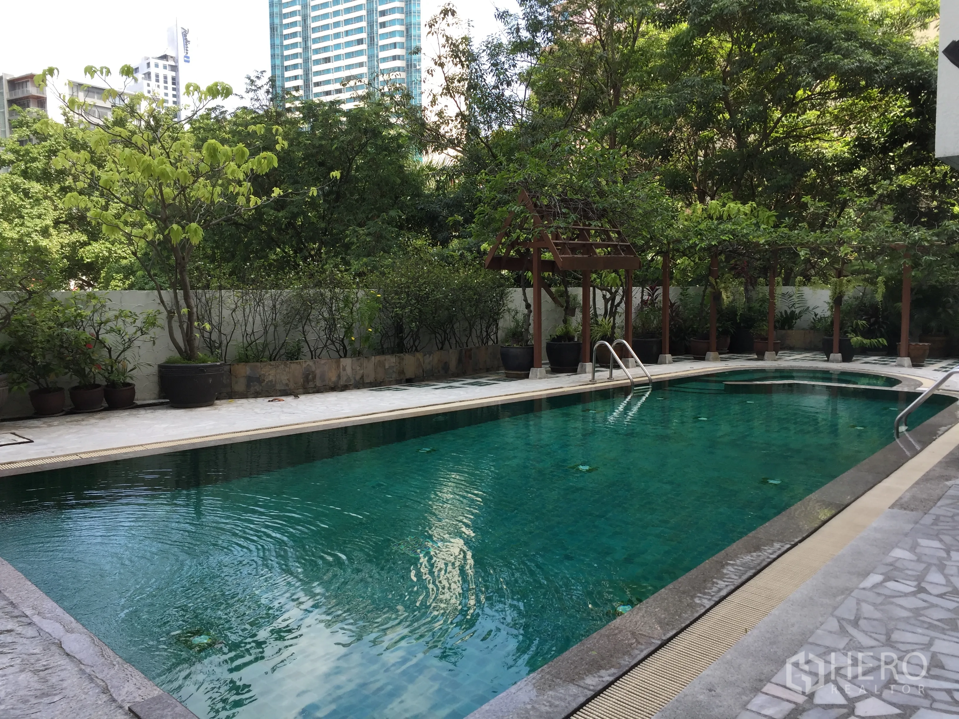 Condominium for rent in Khlong Toei, Bangkok - Outdoor condominium swimming pool surrounded by greenery and wooden pergolas with city buildings in the background.