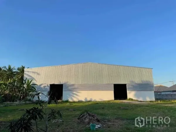 Warehouse for sale in Chon Buri - Large corrugated steel warehouse with multiple wide door openings and a grassy yard under a clear blue sky.