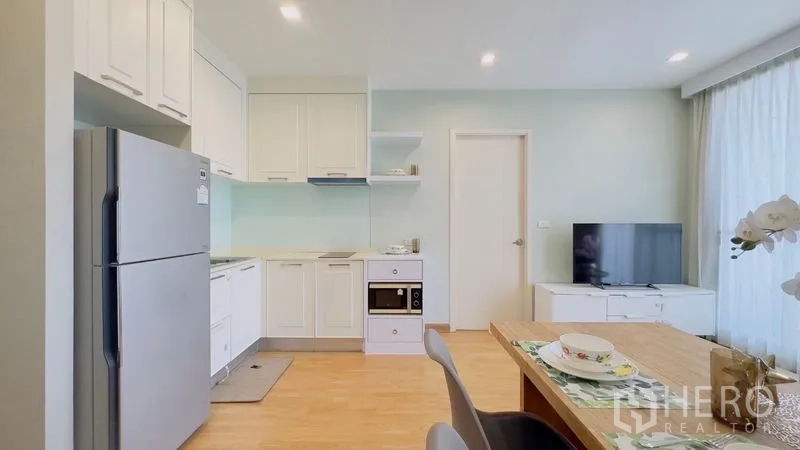 Condominium for rent in Watthana, Bangkok - Compact white kitchen with cabinets, cooktop, oven, stainless refrigerator and TV area.