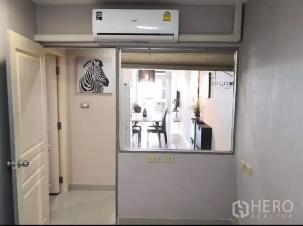Condominium for sale in Khlong Toei, Bangkok - Compact room with wall-mounted air conditioner and interior window looking toward the dining area, neutral gray walls.