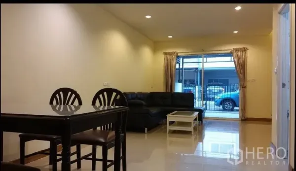 Townhouse for rent in Suan Luang, Bangkok - Bright living and dining area with black leather sofa, dining set, and sliding glass doors opening to a gated patio.