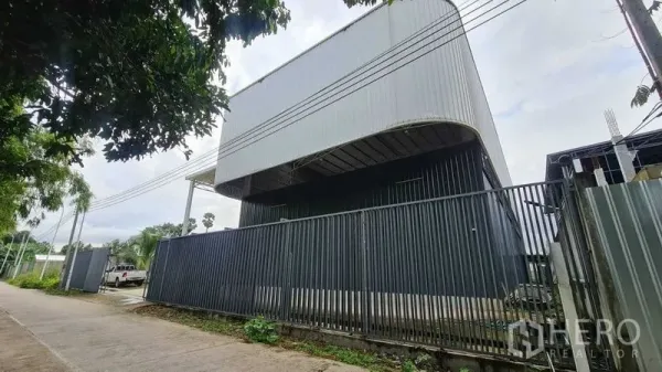 Warehouse for rent in Mueang Nonthaburi, Nonthaburi - Street view of a modern steel-clad warehouse with a curved high roof and tall perimeter fence in Nonthaburi.