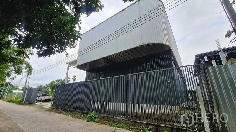 Warehouse for rent in Mueang Nonthaburi, Nonthaburi - Street view of a modern steel-clad warehouse with a curved high roof and tall perimeter fence in Nonthaburi.
