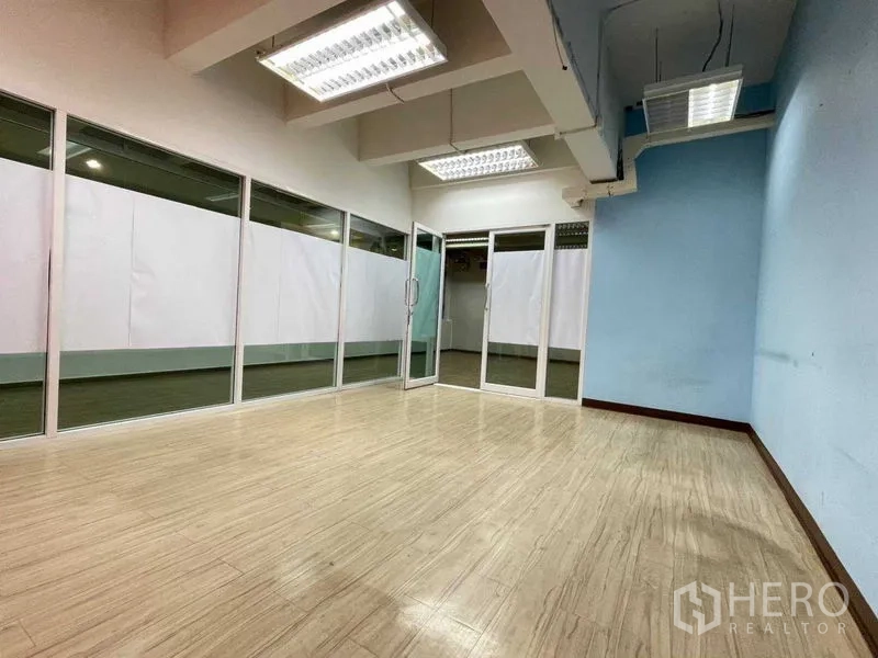 Office Space for rent in Ratchathewi, Bangkok - Spacious empty room with full-height glass wall, pale timber flooring and suspended fluorescent lighting.