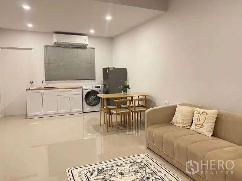 联排别墅 出租在Prawet, Bangkok - 开放式茶水厨房与餐区，含沙发、冰箱、洗衣机与白色橱柜。