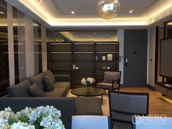 Condominium for rent or sale in Watthana, Bangkok - Modern living room with dark built‑in shelving, grey sofas, round coffee table and cove lighting on wood floors.