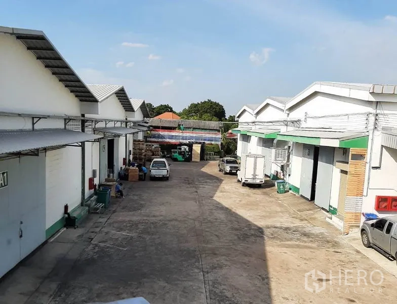 Warehouse for rent in Suan Luang, Bangkok - Row of low-rise warehouses with roll-up doors and trucks in a wide service lane.