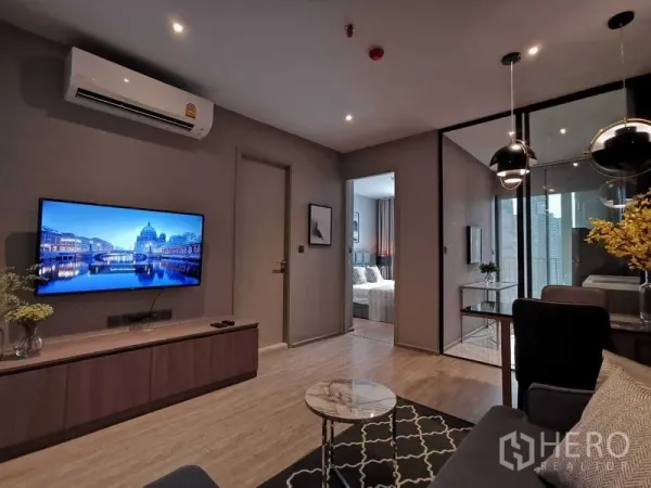Condominium for sale in Watthana, Bangkok - Modern living room with wall-mounted TV, wood flooring, and glass doors leading to the kitchen and city-view balcony.