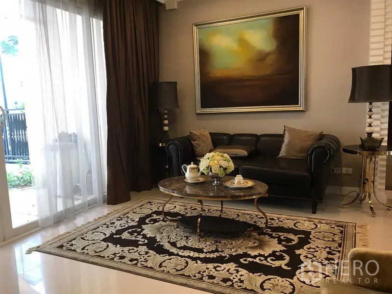 Townhouse for rent or sale in Bang Kho Laem, Bangkok - Elegant living room with leather sofa, abstract artwork, coffee table and patterned rug beside floor‑to‑ceiling sliding glass doors.