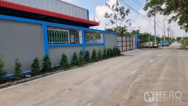 คลังสินค้า ให้เช่าในMin Buri, Bangkok - ด้านข้างอาคารโกดัง กำแพงรั้วยาวมีช่องหน้าต่างเหล็ก ต้นไม้ตกแต่ง และถนนเงียบพร้อมสายไฟฟ้า