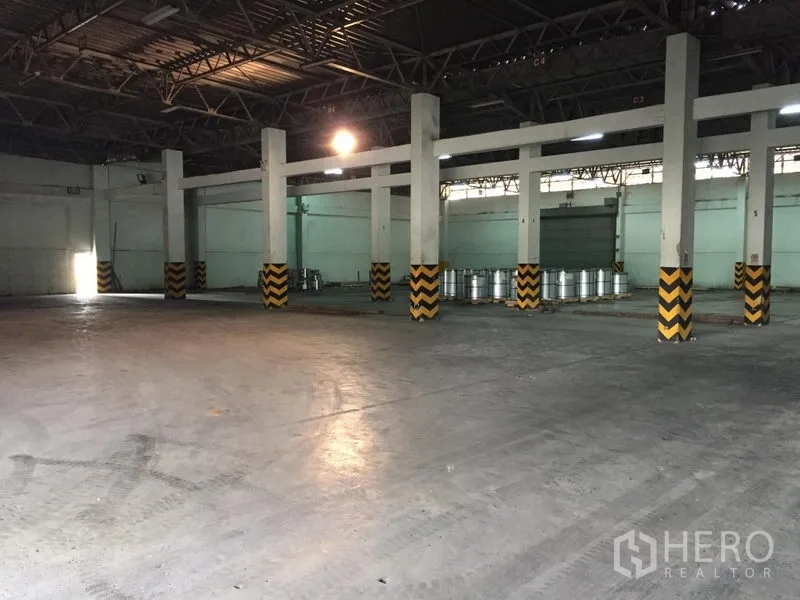 货栈 出租在Pathum Thani - 宽敞仓库内部，高钢结构屋顶、混凝土地面与黄黑警示条立柱。