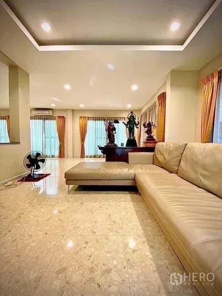 Detached House for sale in Saphan Sung, Bangkok - Bright living room with L‑shaped leather sofa, polished marble flooring, recessed lights and large windows with curtains.