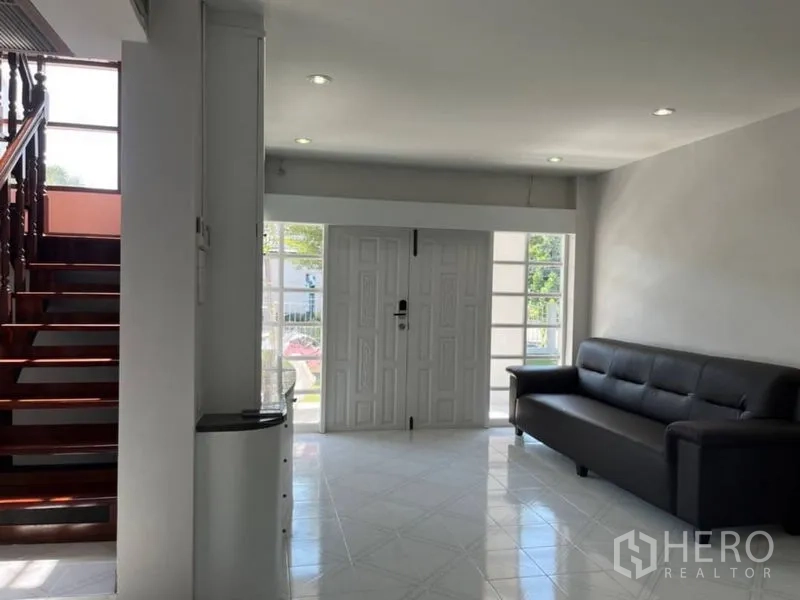 Detached House for rent in Samut Prakan - Light‑filled living area with white double doors, sofa, and wooden staircase.