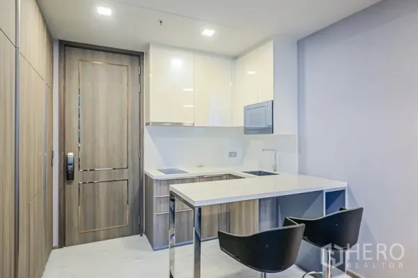 Condominium for rent in Watthana, Bangkok - Modern kitchenette with induction hob, glossy cabinets and a breakfast bar with two stools by the entry.