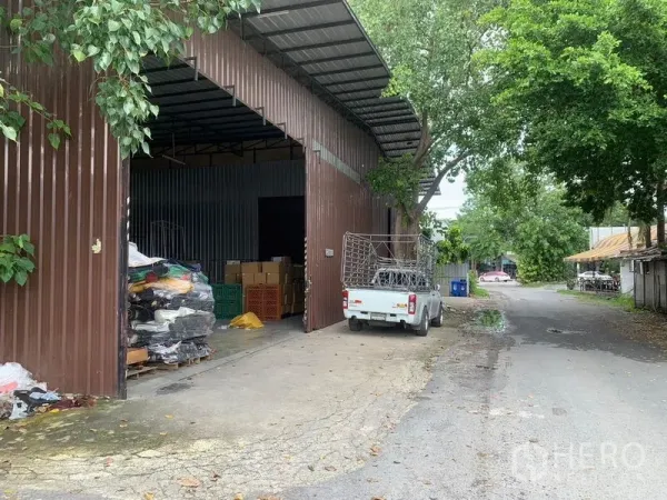 货栈 出租在Bang Kapi, Bangkok - 邦甲比树荫道路旁的金属外墙仓库，宽大推拉门与装卸区，可停皮卡车。