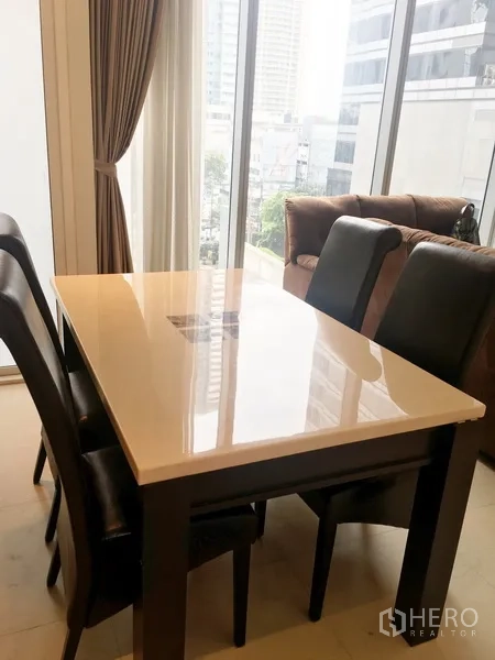 Condominium for rent in Bang Rak, Bangkok - Dining area with a glossy white table and four leather chairs beside tall windows overlooking the city.