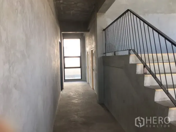 Home Office for rent in Lat Krabang, Bangkok - Industrial-style corridor with raw concrete walls and a metal‑railed staircase inside the building.
