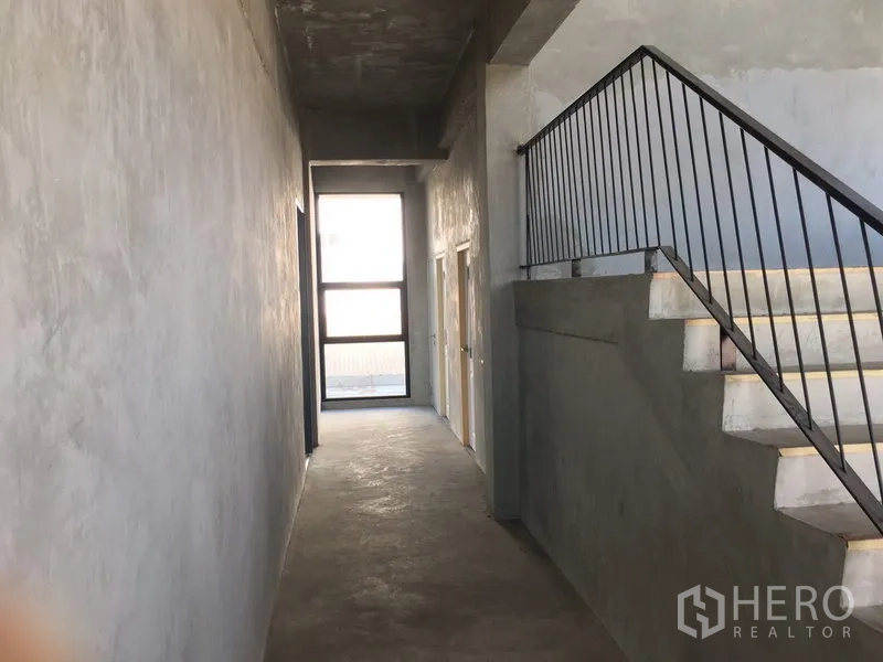 Home Office for rent in Lat Krabang, Bangkok - Industrial-style corridor with raw concrete walls and a metal‑railed staircase inside the building.