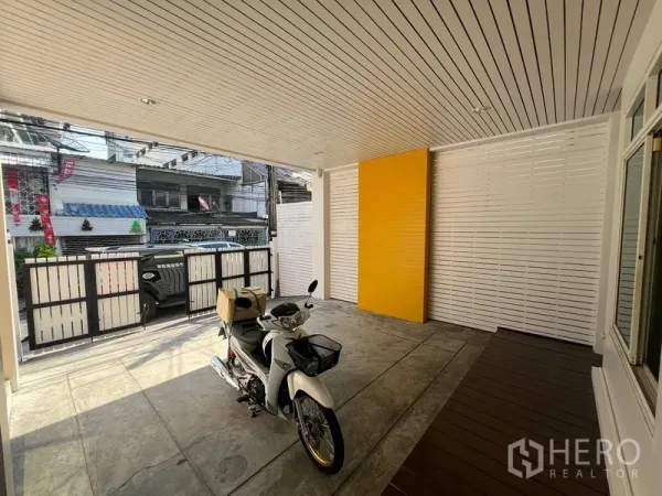Townhouse for rent in Din Daeng, Bangkok - Shaded carport with white slatted ceiling, yellow feature wall and gated entrance on concrete floor.
