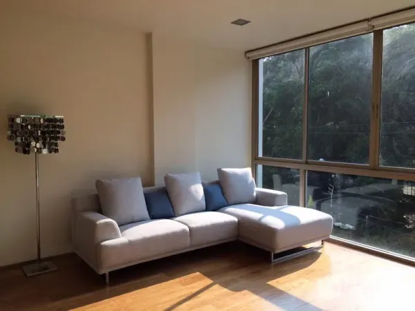 Condominium for rent in Khlong Toei, Bangkok - Bright living room with L-shaped sofa, floor lamp, and large windows overlooking trees.