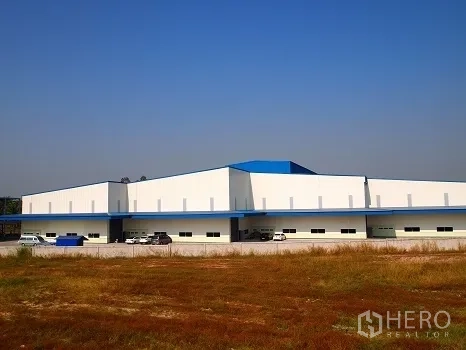 โรงงาน ให้เช่าในSi Racha, Chon Buri - ภาพมุมกว้างตัวอาคารโรงงานชั้นเดียวสีขาวหลังคาสีน้ำเงิน มีที่จอดรถ มองจากพื้นที่โล่ง