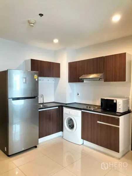 Condominium for rent in Watthana, Bangkok - Compact L-shaped kitchen with wood cabinets, fridge, washing machine and microwave.