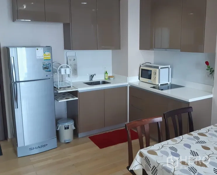 Condominium for rent in Khlong Toei, Bangkok - Compact L-shaped kitchen with glossy cabinets, stainless fridge, microwave and dining table.