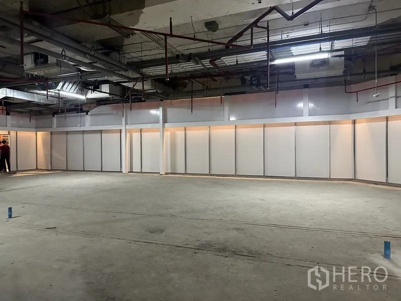 Retail Space for rent - Large rectangular retail area facing a long illuminated partition wall with unfinished flooring.