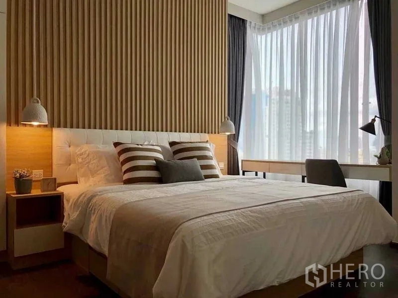 公寓 出租在Watthana, Bangkok - 转角卧室配大床、条纹抱枕、吊灯与书桌，薄纱窗帘映出城市景色。