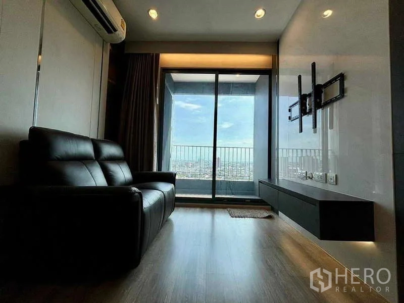 Condominium for rent or sale in Bang Na, Bangkok - Modern living room with black leather sofa, TV wall and sliding glass doors opening to a balcony with city skyline view.