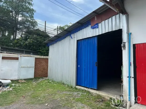 Warehouse for rent in Mueang Samut Prakan, Samut Prakan - Corrugated metal warehouse exterior with blue sliding door beside a brick wall and greenery.