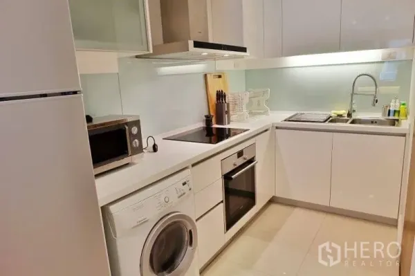 Condominium for rent in Khlong Toei, Bangkok - Modern L-shaped kitchen with oven, induction hob, extractor hood, sink and washing machine.