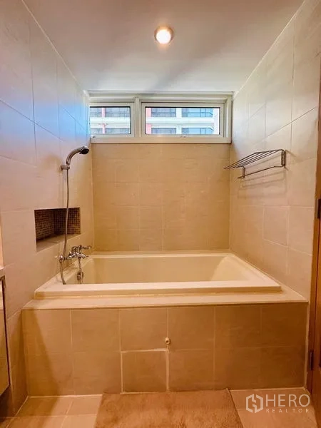 Condominium for rent in Bang Rak, Bangkok - Deep soaking bathtub with handheld shower beneath a high window and tiled walls.