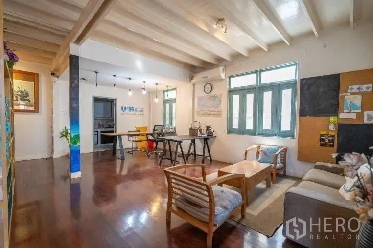 Detached House for rent in Khlong Toei, Bangkok - Open-plan living and work area with wooden floors, beamed ceiling, sofa set, and large windows.
