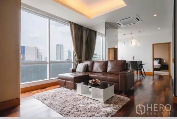 公寓 出租在Sathon, Bangkok - 采光充足的客厅，落地窗、棕色皮沙发、灯槽天花、木地板与城市景观。