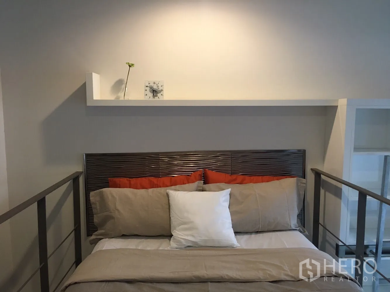 Condominium for rent in Khlong Toei, Bangkok - Loft bedroom with shelf and clock above the headboard, warm neutral tones and safety railings.
