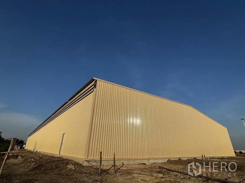 Warehouse for rent or sale in Pak Kret, Nonthaburi - Exterior side view of beige corrugated metal warehouse under a clear blue sky.
