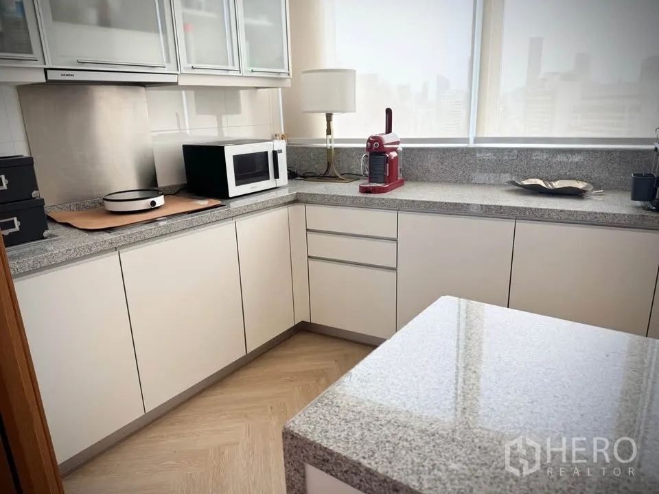 Condominium for rent or sale in Khlong Toei, Bangkok - L-shaped kitchen with granite island and countertops lit by daylight through blinds.