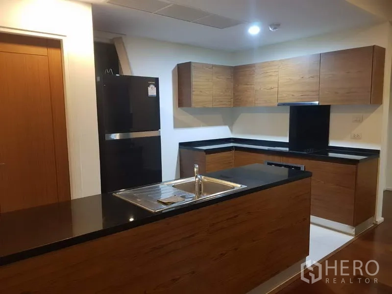 公寓 出租在Watthana, Bangkok - 现代L形厨房，木质柜体与黑色石材台面，带水槽中岛和大冰箱。